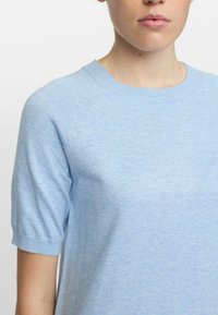 Světle modrý top s krátkým rukávem vyrobený z jemného pleteného materiálu. Má kruhový výstřih a design rukávů raglan. Hladká textura s jemným pružením.