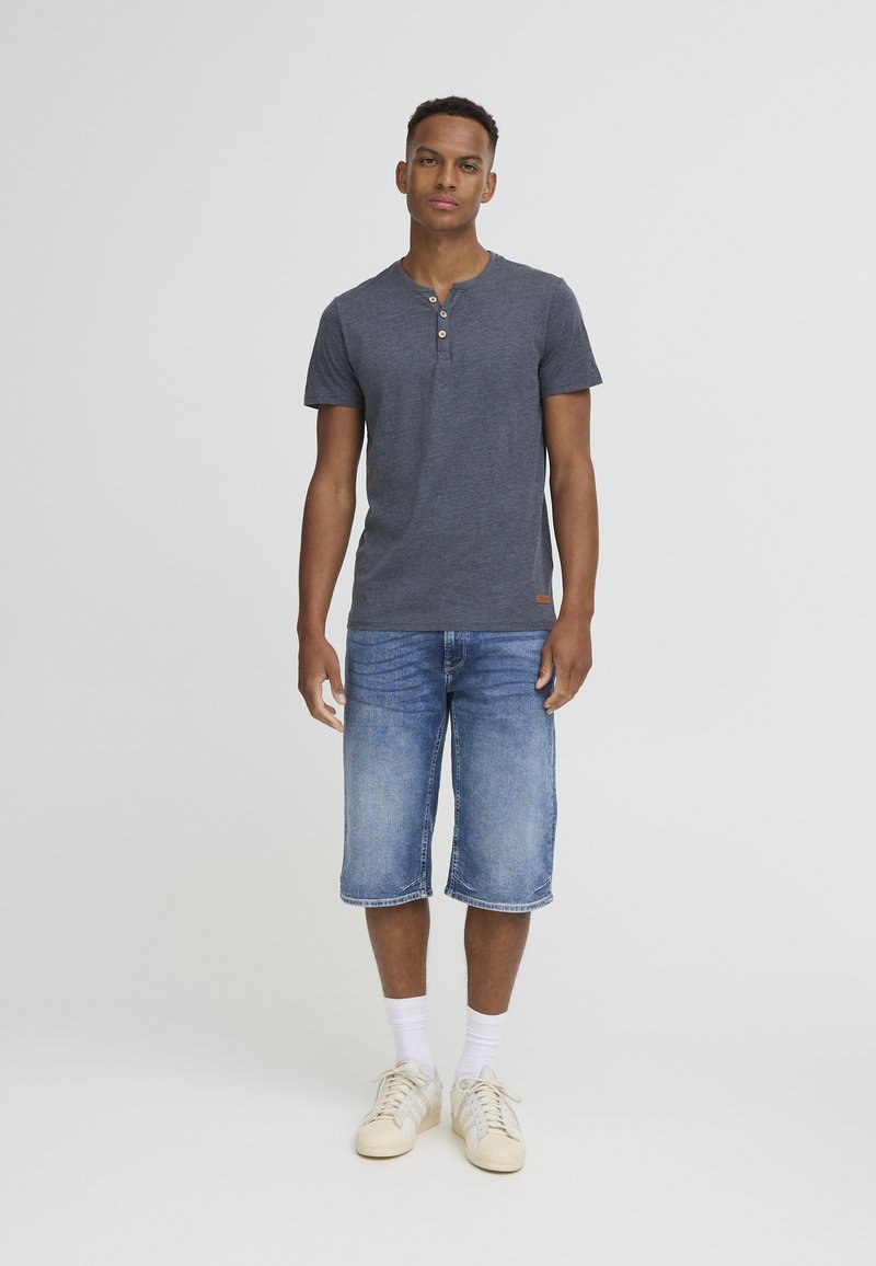 Camiseta henley de manga corta en gris oscuro, con abertura de tres botones, combinada con pantalones cortos de mezclilla azul claro y zapatillas blancas. Tejido liso y texturizado.