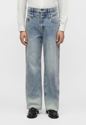 Blugi bleu deschis cu picior larg și detalii la cusatura laterală, purtați cu o cămașă albă cu mâneci lungi și pantofi negri, arătați de la talie până la picioare.