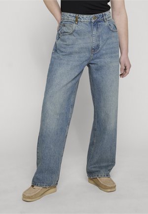 Personne portant un jean en denim bleu clair à coupe ample et des chaussures beige à enfiler, debout avec une main dans la poche sur un fond uni.