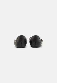Schwarze Lederloafers mit glatter Textur und runder Zehenpartie. Sie haben einen goldenen Akzent und eine flache Sohle und sind von hinten betrachtet.