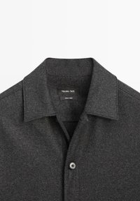 Ciemnoszara koszula z kołnierzykiem wykonana z miękkiego materiału. Posiada zapięcie na guziki oraz metkę z napisem „Massimo Dutti”, wskazującą markę i pochodzenie.