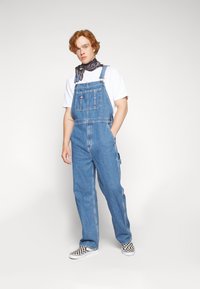 Középkék farmer overál, állítható pántokkal és elülső zsebbel. Fehér pólóval és kockás slip-on cipőkkel párosítva.