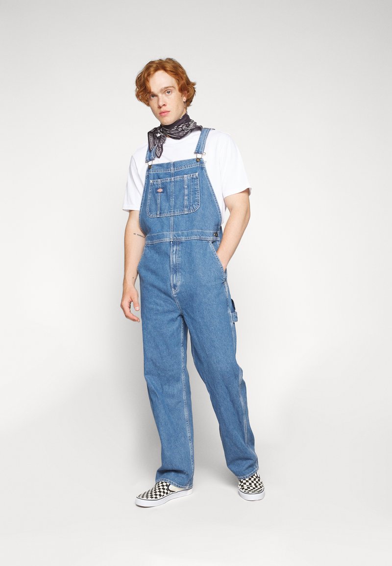 Középkék farmer overál, állítható pántokkal és elülső zsebbel. Fehér pólóval és kockás slip-on cipőkkel párosítva.