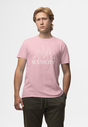 Junger Mann mit blondem Haar, der ein pinkes T-Shirt mit einer weißen Stadtsilhouette und dem Text "SEX AND THE CITY" sowie dunkelgrüne Hose trägt.