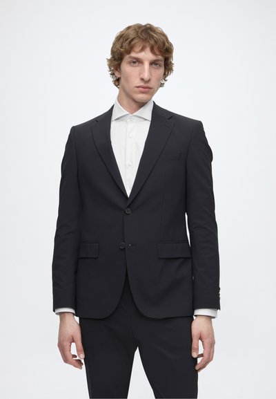 Jeune homme aux cheveux clairs et bouclés, portant un costume noir et une chemise blanche, debout devant un fond blanc uni.