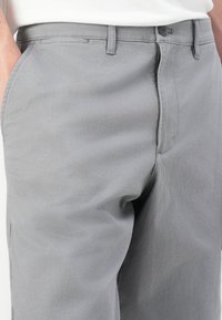 Gros plan sur une personne portant un pantalon gris clair avec des poches latérales, des passants de ceinture et une fermeture à bouton, les mains partiellement dans les poches.