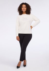 Felpa crema con "Cozy Season" ricamato in grigio, abbinata a pantaloni neri e tacchi neri. Tessuto liscio con polsini a costine.