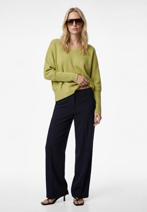 Kvinde iført oversized lysegrøn sweater, sorte bukser med vide ben, sorte sandaletter med hæl, solbriller og sølvringe, poserer mod hvid baggrund.