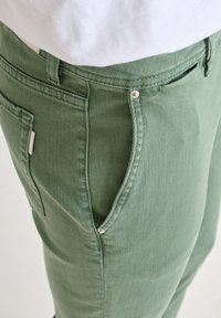 Pantalones de algodón verde con textura de denim, con bolsillos laterales y acentos de metal. La vista cercana resalta los detalles de las costuras.