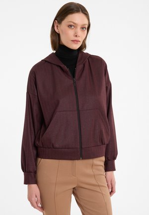 Vrouw in een bordeauxrood ritsvest met verticale strepen over een zwarte coltrui en beige broek, staande tegen een witte achtergrond.