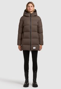 Frau in brauner, mit Kapuze versehenem Steppjacke, schwarzer Leggings und schwarzen Stiefeln, die vor einem schlichten, hellen Hintergrund steht.