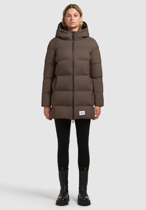 Vrouw in een bruin gewatteerd jack met capuchon, zwarte leggings en zwarte laarzen, staand tegen een effen lichte achtergrond.