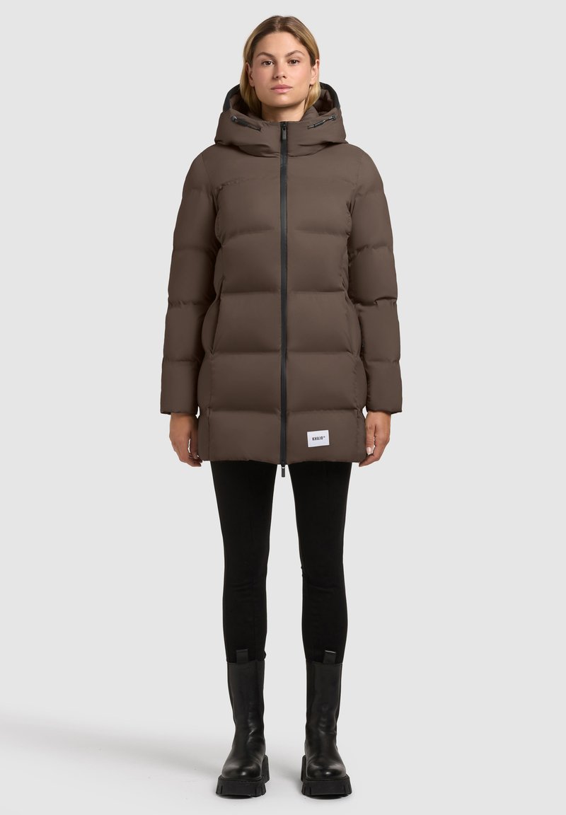 Frau in brauner, mit Kapuze versehenem Steppjacke, schwarzer Leggings und schwarzen Stiefeln, die vor einem schlichten, hellen Hintergrund steht.