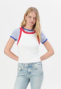 Hvit ribbet tank topp med røde detaljer og blå stripete korte ermer. Kombinert med lyseblå jeans, som viser en avslappet passform.