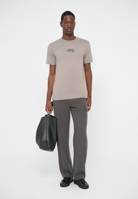 Beige cotton t-shirt with small black text, paired with dark grey trousers and a black leather bag. Black shoes complete the outfit.