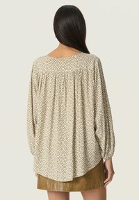 Blusa beige a pois con maniche lunghe, yoke posteriore arricciato e orlo arrotondato. La texture appare morbida e leggera.