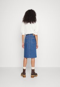 Mujer con cabello rizado, vestida con una blusa blanca texturizada, una falda de mezclilla azul hasta la rodilla, calcetines beiges y botas de tacón marrones, de pie con la espalda a la cámara.