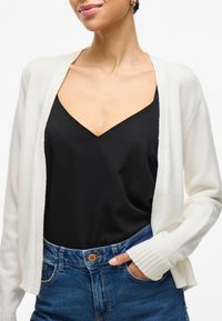 Cardigan bianco aperto davanti con polsini a coste, indossato sopra un top nero a scollo a V. L'outfit è abbinato a jeans in denim blu a vita alta.