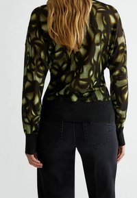 Tumma kuvioitu neule, jossa on vihreä kukkakuosi mustalla pohjalla, istuvalla mustalla vyötärönauhalla ja pitkät hihat.