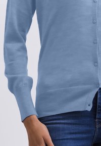 Närbild av en person som bär en ljusblå knappad cardigan och mörkblå jeans, med sin vänstra arm avslappnat vid sidan.