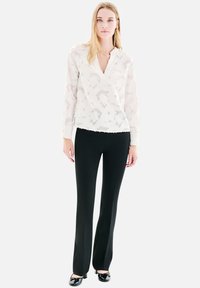 Femme debout portant un blouse long à manches blanches à motifs, un pantalon noir droit et des chaussures vernies noires à petits talons.