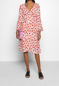 Eine Frau trägt ein weißes Wickelkleid mit roten Herzmustern, hält eine pinke Clutch und schwarze offene Riemchensandalen vor einem schlichten Hintergrund.