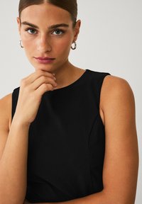 Sort ærmeløs kjole med rund halsudskæring og glat tekstur; modellen har hånden nær sin hage og viser små hoopsøreringe.