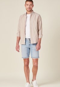 Ljust beige långärmad skjorta över en vit t-shirt, i kombination med ljusblå denimshorts och vita sneakers. Avslappnad stil, ledig passform.