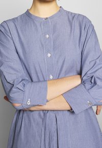 Modro-bílý pruhovaný shirt s kulatým límečkem, zapínáním na knoflíky vpředu a měkkou tkaninou; má dlouhé rukávy a manžety na knoflíky.