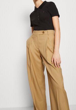 Henkilö, jolla on musta lyhythihainen poolopaita ja korkeavyötäröiset, leveälahkeiset beigehousut, joissa on napitukset.