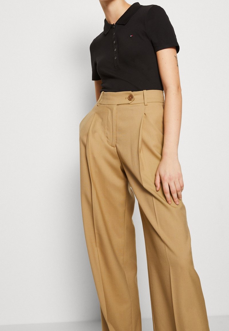 Personne portant un polo noir à manches courtes et un pantalon beige taille haute à jambes larges avec une fermeture à bouton.