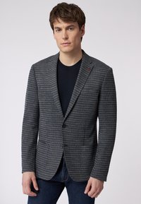 Grahamuster-Blazer mit Reverskragen, zwei Knöpfen, Fronttaschen und einem dezenten roten Akzent am Kragen. Getragen über einem schwarzen Hemd.