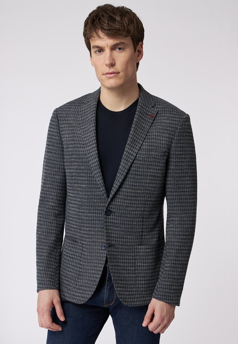 Grahamuster-Blazer mit Reverskragen, zwei Knöpfen, Fronttaschen und einem dezenten roten Akzent am Kragen. Getragen über einem schwarzen Hemd.