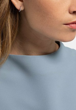 Nahaufnahme des unteren Gesichts und des Nackens einer Frau, die einen kleinen silbernen Creolenohrring und ein hellblaues, strukturiertes Oberteil trägt.