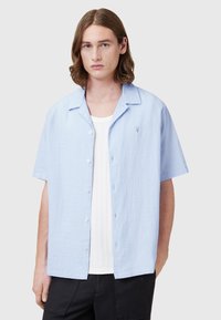 Lichtblauw shirt met korte mouwen van een gestructureerde stof, knoopsluiting en een subtiele logo op de linkerborst, gelaagd over een witte top.