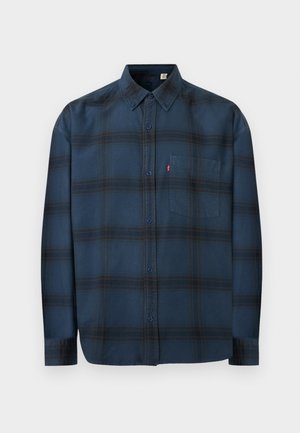 Chemise bleu foncé à manches longues avec motif à carreaux horizontal noir, col boutonné, poche poitrine et étiquette rouge sur la poche.
