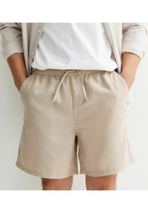 Persona che indossa pantaloncini beige con coulisse, t-shirt bianca e camicia a righe marrone chiaro con bottoni, con le mani in tasca.