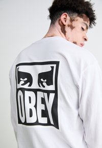 Long-sleeve white t-shirt featuring a large black graphic design on the back, displaying stylized eyes above the word "OBEY."