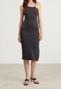 Černé, přiléhavé midi šaty vyrobené z hladké tkaniny s tenkými ramínky, končící těsně pod kolenem, doplněné o černé sandály na podpatku.