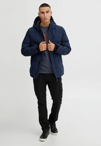 Chaqueta de plumas azul marino con capucha, cremallera frontal y detalles en los puños, llevada sobre una camiseta gris y combinada con pantalones cargo negros y zapatillas.