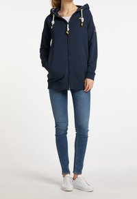 Marin blå zip-up hoodie med huva, snörningar i gult och vitt, avslappnad passform, matchad med blå skinny jeans och vita sneakers.