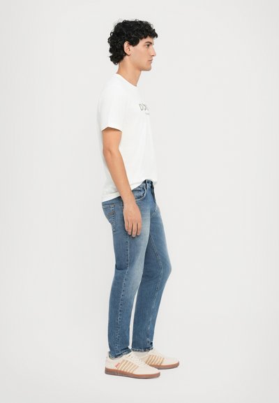 T-shirt bianca in cotone con logo, jeans in denim blu e sneakers beige con accenti arancioni e strisce. Indossato da una persona di profilo.