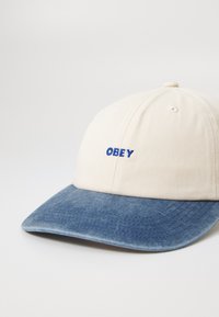 Creme baseball cap med skygge i blå denim, med "OBEY" broderet i blå på forsiden. Glat stof, struktureret form.