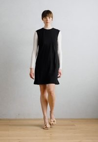 Zwarte mouwloze jurk met een losse pasvorm, gemaakt van lichtgewicht stof. Gecombineerd met naakte schoenen met een rimpelaccent. Eenvoudig en elegant ontwerp.