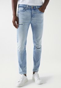 Vaqueros de mezclilla azul claro con parches desgastados en las rodillas, diseño de cinco bolsillos y corte recto, combinados con zapatillas blancas.