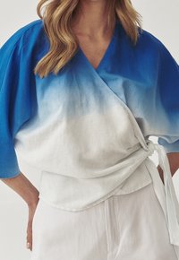 Top envolvente en degradado azul y blanco, hecho de un tejido ligero, con mangas anchas y detalle de cintura anudada. Textura suave, corte relajado.