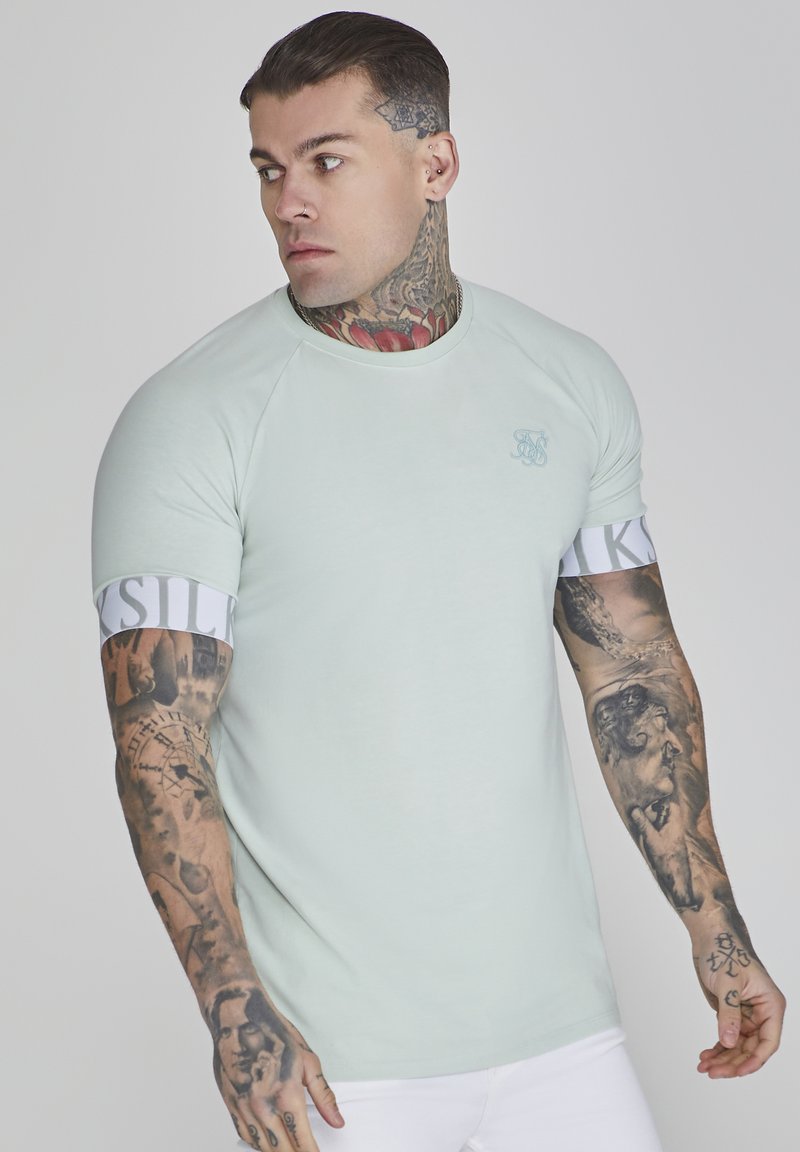 T-shirt fitted verde chiaro con maniche raglan, caratterizzato da una fascia bianca con il testo "SILK". Il design include un marchio discreto e tatuaggi sulle braccia.