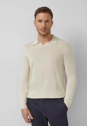 Mężczyzna z krótkimi brązowymi włosami, ubrany w kremowy sweter z długim rękawem i kołnierzykiem oraz ciemne spodnie, stoi na jasnym, jednolitym tle.