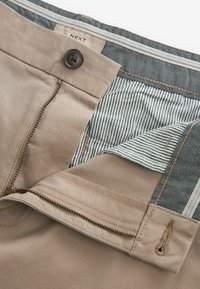 Bež hlače s patentnim zatvaračem, kopčanjem na gumbe i podstavom od prugastog materijala. Imaju glatku teksturu i kontrastne šavove.